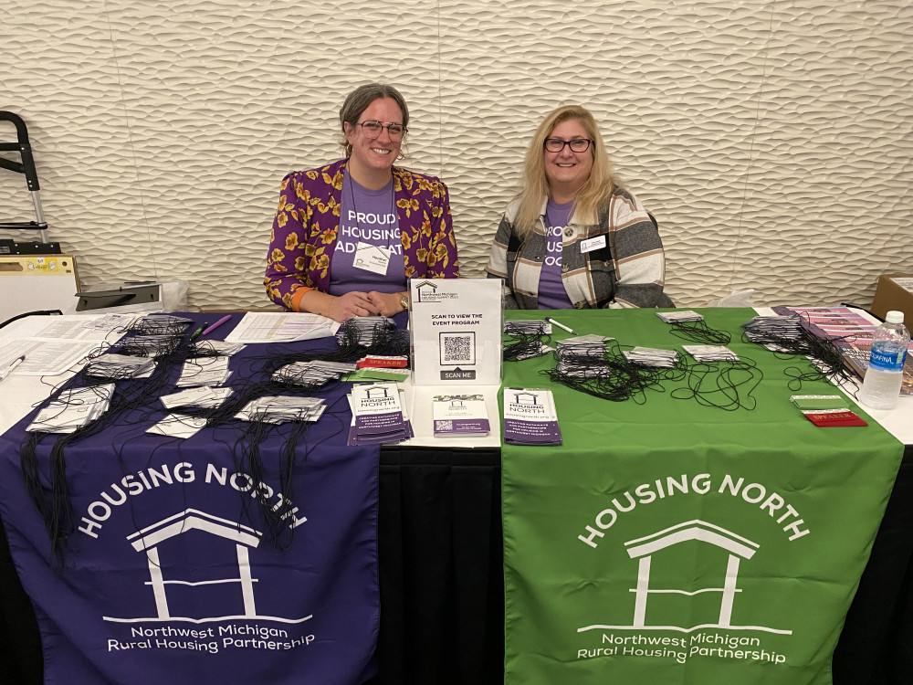 Two Housing North staff members sitting at a Housing North registration table with name tags and pamphlets on it.