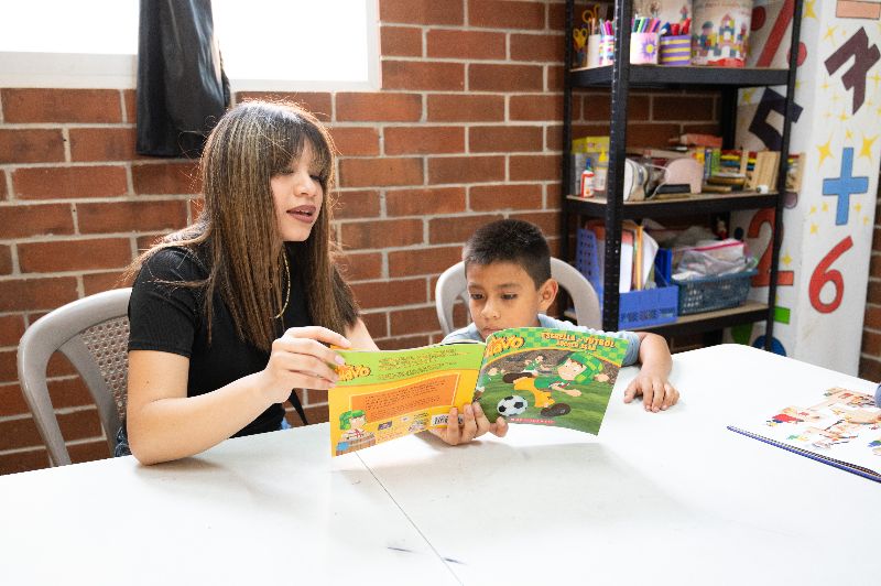 Image description: A young woman reads a colorful children's book to a young boy in a classroom setting. The background features books, educational materials, and vibrant decor.