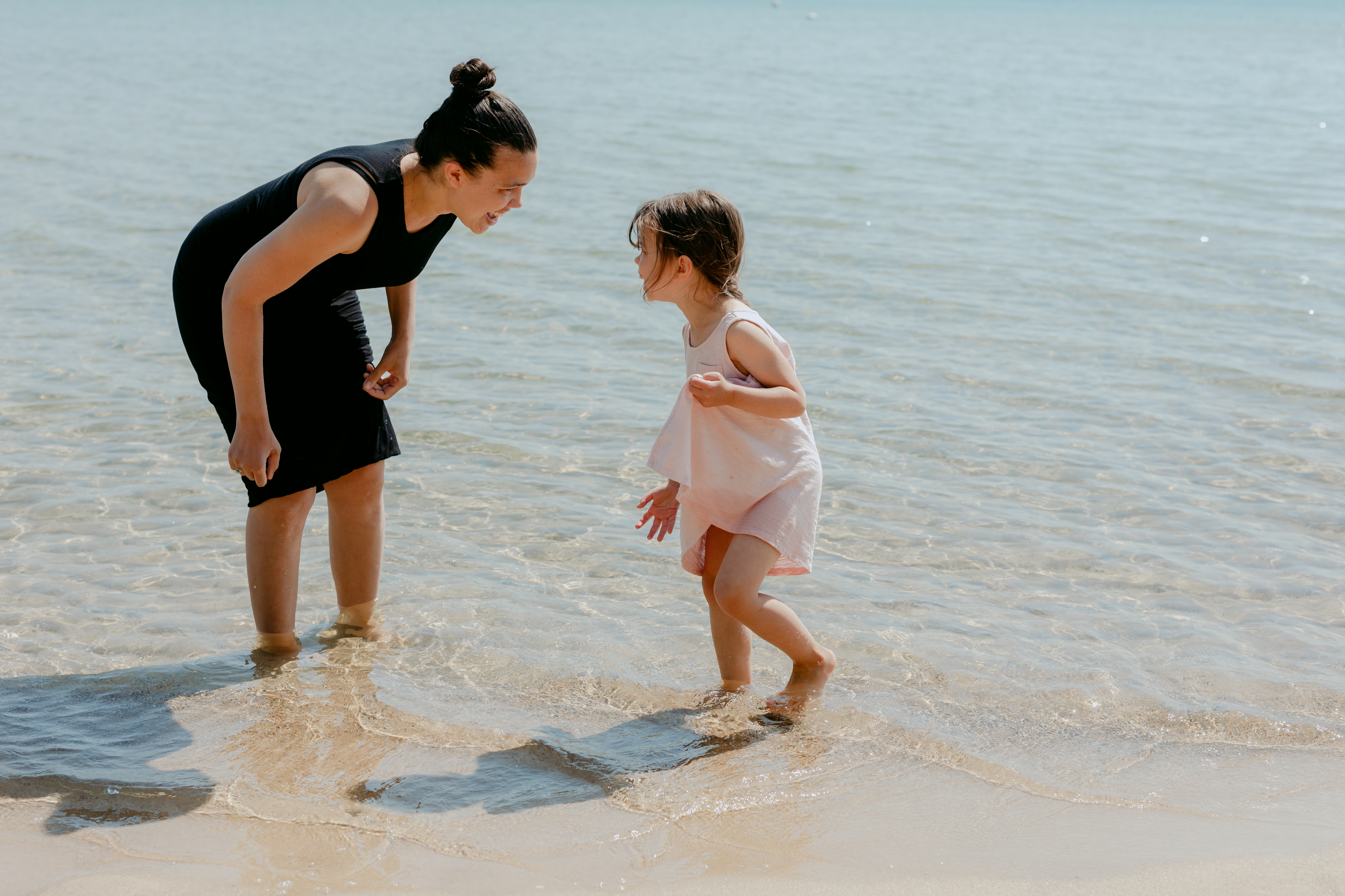 A mother and her young daughter laugh and play together in shallow water at the beach on a sunny day.