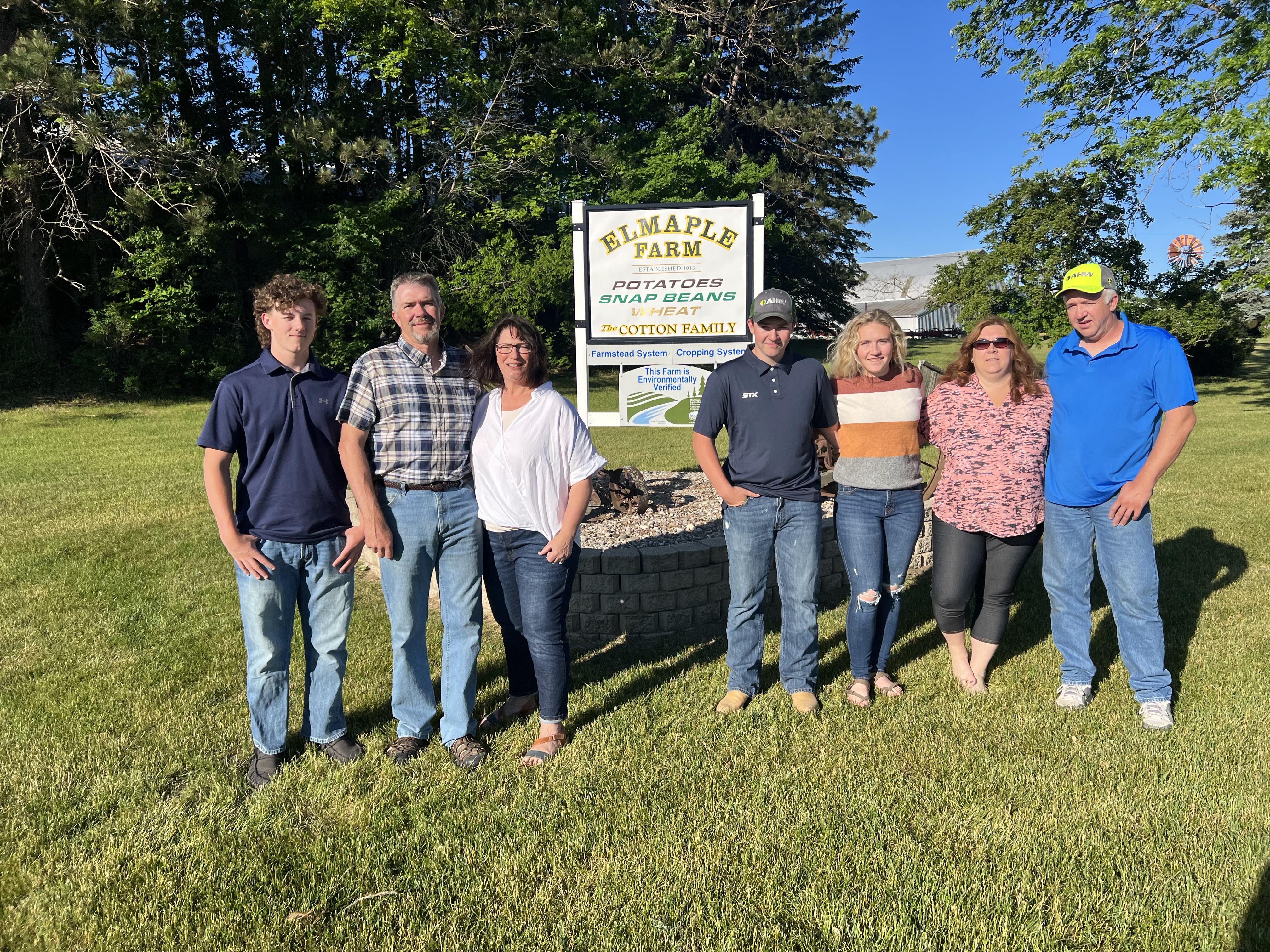 The Elmaple Farm Family standing together in front of the farm sign outside on a bright day